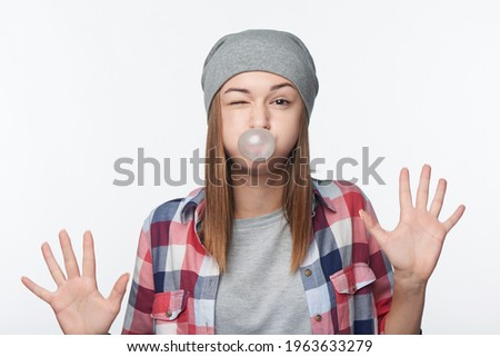 Fooling teen girl blowing bubblegum showing palms, winking, studio portrait over white background