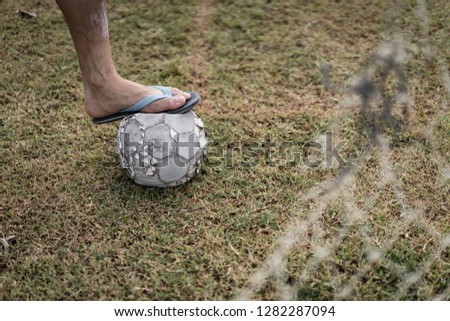 The foot of football player put on the old football in the grass field with the foreground of broken old goal net. Concept of people and sport in poor country.