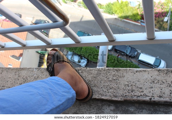Foot in sandals on the 10th floor balcony with\
cars underneath