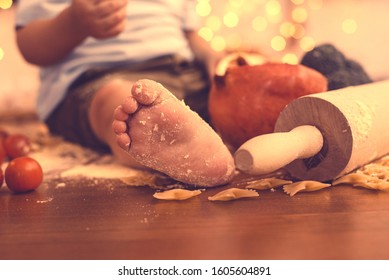 The Foot Of A Young Child In The Kitchen. The Concept Of Baby Food, Care For Children And Their Nutrition. Helping A Child Mom With Cooking, Pasta And Salads.