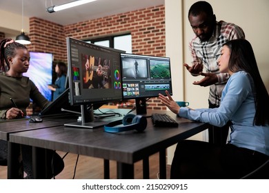 Footage editing specialist using multi monitor workstation to improve frames quality. Professional videographer sitting at desk while coworker helping her with film visual improvement.
