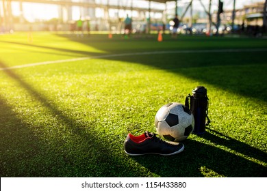 Football, cool flask and black red sports shoes on green artificial turf with blurry kid soccer player training. Sunshine and shadows of soccer training equipment. - Powered by Shutterstock