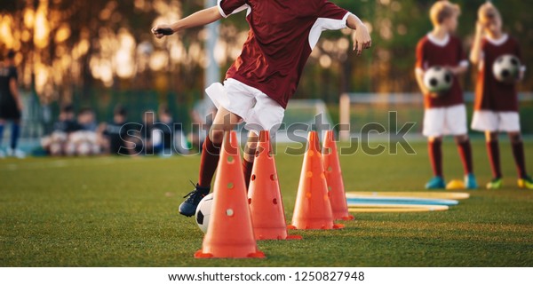 Football Drills: The Slalom Drill. Youth soccer\
practice drills. Young football players training on pitch. Soccer\
slalom cone drill. Boy in red soccer jersey shirt running with ball\
between cones