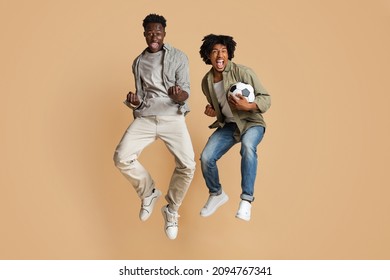 Football Fans. Full Length Shot Of Two Euphoric Black Guys Jumping In Air With Soccer Ball In Hands, Excited African American Friends Emotionally Reacting To Win, Having Fun On Beige Background