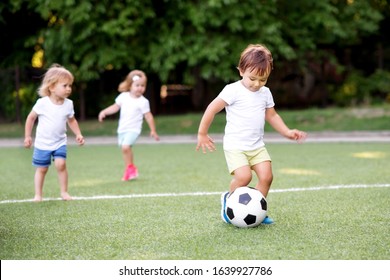 Football Game: Team Of Toddlers Playing Soccer On Green Field: Three Kids, Two Boys (one Is Barefoot) And Girl Playing At Stadium, Smiling Little Boy Dribbling Ball Is Running In Front Place