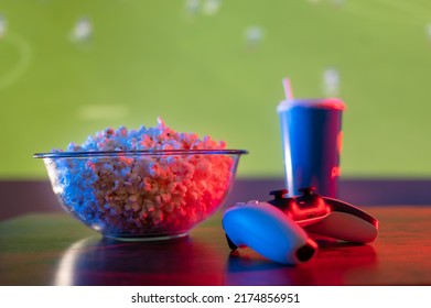 Football Match On The Big TV Screen. On A Wooden Table Are Popcorn In A Bowl, A Carbonated Drink In A Plastic Glass And A Joystick. Video Games, Online Games With Friends, Sports.