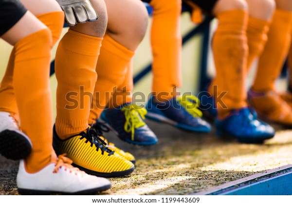 Footballers in soccer\
cleats. Youth athletes in soccer clothes. Young football players\
wearing football clothes and soccer shoes sitting on bench in a\
row. Soccer detail\
background.