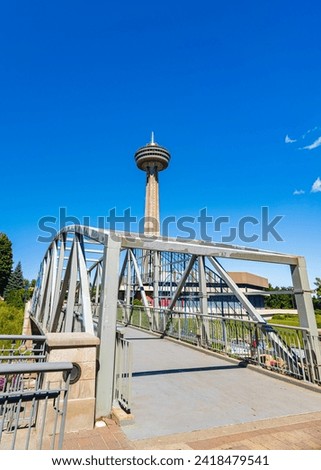 A footbridge in Niagara Park leads towards the iconic Skylon Tower under a clear blue sky in Niagara Falls, Ontario, showcasing architectural and natural harmony, Canada. High quality photo