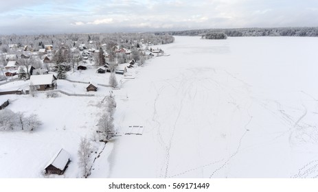 Footprints are on ice of frozen lake near winter shore. Russian remote village is in evergreen forests. Aerial view. Russia