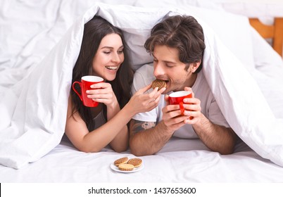 Forbidden snack. Happy millennial couple enjoying cookies and coffee, hiding under duvet in bedroom