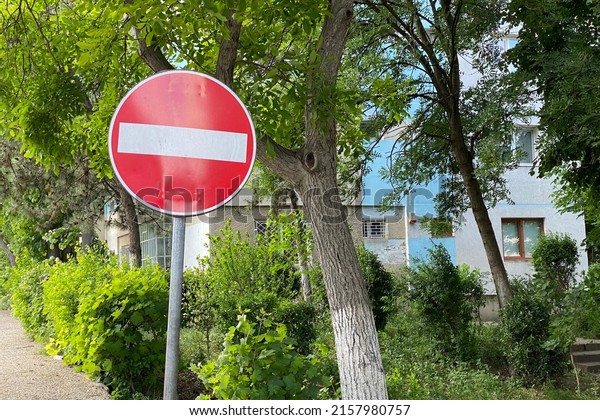 Forbidden traffic\
sign post in red and white with green spring foliage and blue\
building walls in the\
background.