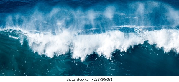 A forceful, crashing wave of the ocean is captured in this stunning aerial photo, with the massive wave breaking into a white spray against the deep waters below, evoking feelings of awe and power. - Shutterstock ID 2289306391