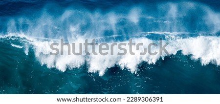 A forceful, crashing wave of the ocean is captured in this stunning aerial photo, with the massive wave breaking into a white spray against the deep waters below, evoking feelings of awe and power.
