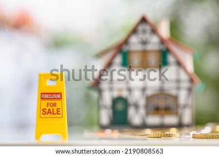 Foreclosures and foreclosed home for sale property listings, financial concept : Yellow warning sign board with the words FORECLOSURE FOR SALE, a two-story half-timbered model house, coins on a table.