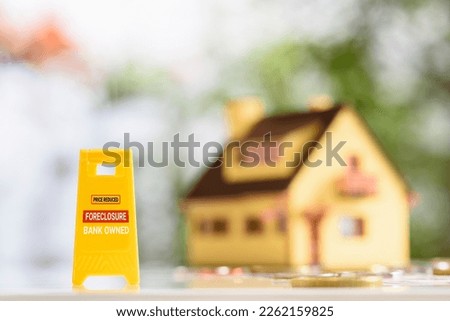 Foreclosures and foreclosed home for sale property listings, financial concept : Yellow warning sign board with the words FORECLOSURE FOR SALE BANK-OWNED, a two-story model house, coins on a table.