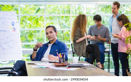 Foreground Focus Handsome Hipster Caucasian Male Creative Director, Boss Or Leader Sitting In Comfortable Modern Office, Smiling With Confidence And Profession With Background Of Mixed Race Employee