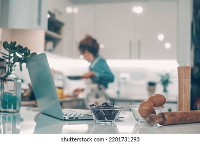 Foreground Focus On Laptop And Food Ingredients In The Kitchen At Home. Woman In Background Defocused Cooking And Enjoying Lunch Preparation Alone. Real Life Lifestyle And Modern Online Web Contents