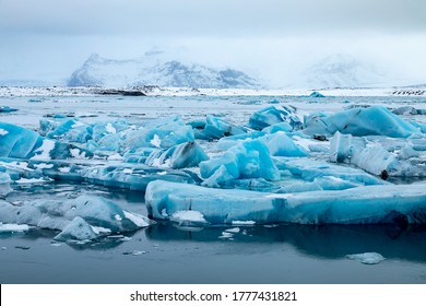 Foreground focus on the rich turquoise blue icebergs floating in the lagoon, which the snow capped mountains int he background. 