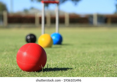 In the foreground a red croquet ball is in close focus on a croquet lawn while the blue, yellow and black balls and the croquet hoop are out of focus in the background