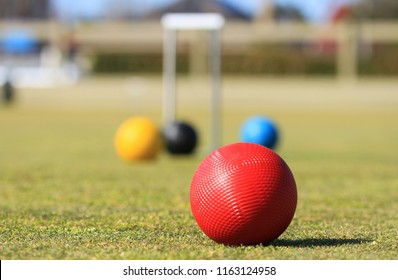 In the foreground is a red croquet ball on a green croquet lawn in Australia with a defocused croquet hoop and balls in the background