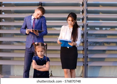 In  foreground stands and frowns, angry with arms crossed in front of little girl, child,  in background successful young handsome man's man, young father and modern woman, young mother hold gadgets