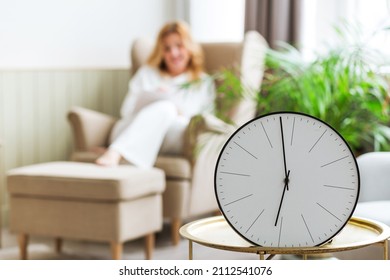 In the foreground there is a clock on a table, in the background a woman is sitting. The concept of unhurried life and relaxation at home