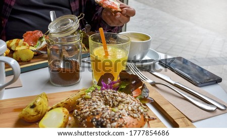 Foreground view of a tabletop with a man hand who are having breakfast, brunch, lunch, or supper next to a window, with coffee, orange juice, cutlery and food.