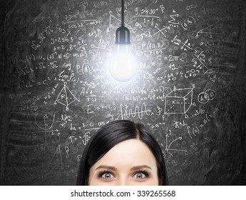 A Forehead Of Brunette Woman Who Is Thinking About Solution Of Complicated Math Problem. Math Formulas Are On The Black Chalkboard. A Light Bulb As A Concept Of A Solution. Analytics Approach.