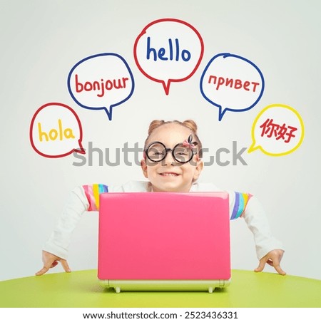 Foreign language learning concept of a beautiful cute little girl with eyeglasses behind a laptop on table. Hello word in Spanish, French, Russian and Chinese. 