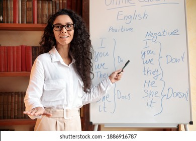 Foreign Language. Portrait of confident smiling young female lecturer wearing glasses teaching English pointing at grammar rules on board with marker, looking back at students at classroom - Shutterstock ID 1886916787