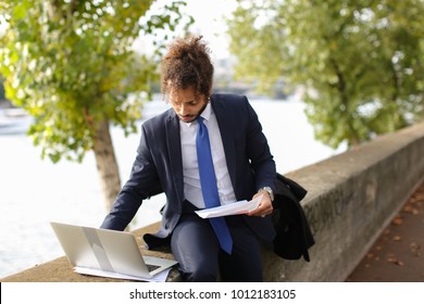 Foreign student writing course work on Seine bank with laptop and papers. Arabian man dressed in suit with tie has long fleecy hair and charming smile. Concept working with documents on open air and