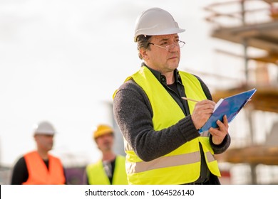 Foreman At Work On Construction Site Checking His Notes And Drawing Plan On Clipboard