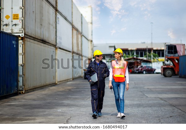 Foreman and worker staff wearing a yellow helmet\
walking in a industrial shipping yard. He\'s consulting on exporting\
goods overseas. Container transportation and export with commercial\
docks.