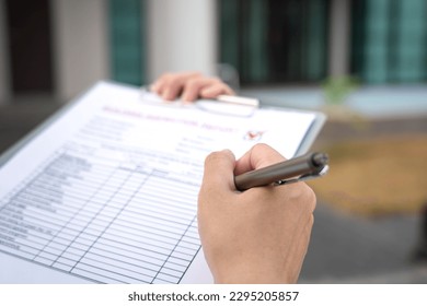 Foreman's hand is using a pen checking on building inspection report form to QC building quality of the house (background). Construction industrial working scene concept. Close-up and selective focus.