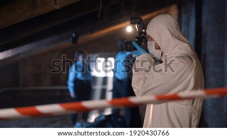 Forensic specialist with photo camera standing at crime scene. Portrait of policeman in protective overall collecting evidence of murder. Police investigation