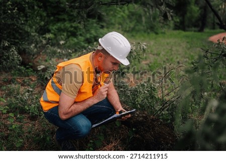 Forest engineer crouching and taking notes on clipboard while inspecting a damaged tree after a storm in a wooded area