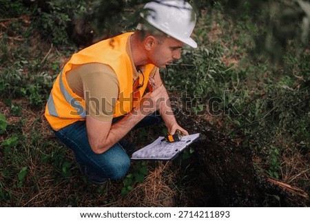 Forest engineer crouching, writing notes on clipboard and measuring a fallen tree trunk using a telemeter