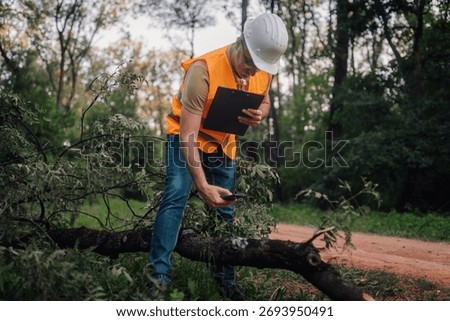 Forest engineer taking notes with clipboard and using mobile phone while inspecting a fallen tree after a storm