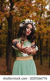 Forest Fairy In A Long Light Green Dress And With A Wreath Of Flowers On Her Head In The Autumn Forest. Forest Nymph In Halloween.