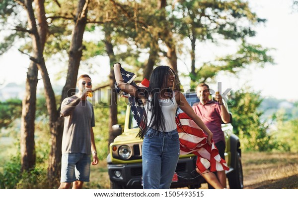 In the forest. Friends have nice\
weekend outdoors near theirs green car with USA\
flag.