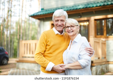 Forest House Of Grandparents: Cheerful Excited Senior Couple Embracing Each Other And Laughing While Looking At Camera And Standing Against Cottage