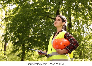 Forester with hard hat and clipboard examining plants in forest, space for text - Powered by Shutterstock