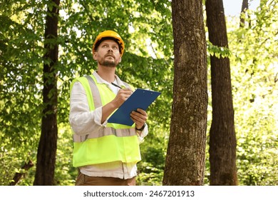 Forester in hard hat with clipboard examining plants in forest - Powered by Shutterstock