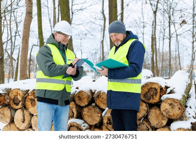 Forestry workers standing in front of a large stack of felled trees in a winter forest. Sale firewood for heating. Two men discussing the purchase dry logs orders during the cold season.	