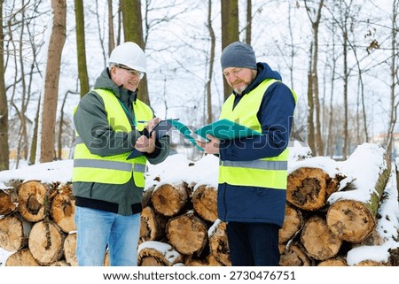 Forestry workers standing in front of a large stack of felled trees in a winter forest. Sale firewood for heating. Two men discussing the purchase dry logs orders during the cold season.	
