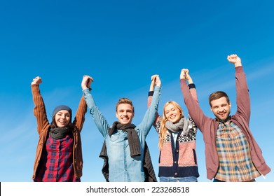 Forever Young. Low Angle View Of Four Young Happy People Holding Hands And Raising Them Up With Blue Sky As Background