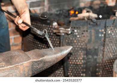 A forger wearing blue jeans and a t-shirt holds fire thongs and a metal twisted bolt on a vintage anvil. There's rounding hammer tool with a wooden handle in the blacksmith foundry shop with a furnace - Powered by Shutterstock