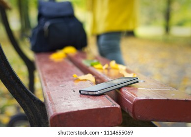 Forgotten Smartphone On A Park Bench. Woman Is Leaving From A Bench Where She Lost Her Cell Phone.