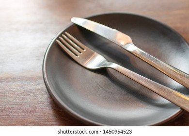 Fork and knife on a black empty plate, wooden table with nature light.