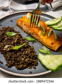 Fork Scooping A Baked Honey-soy Glazed Salmon Fish Fillet Served With Black Beluga Lentils, Cucumber And Green Onions. Healthy Fresh Salmon For Lunch Or Dinner. Side View